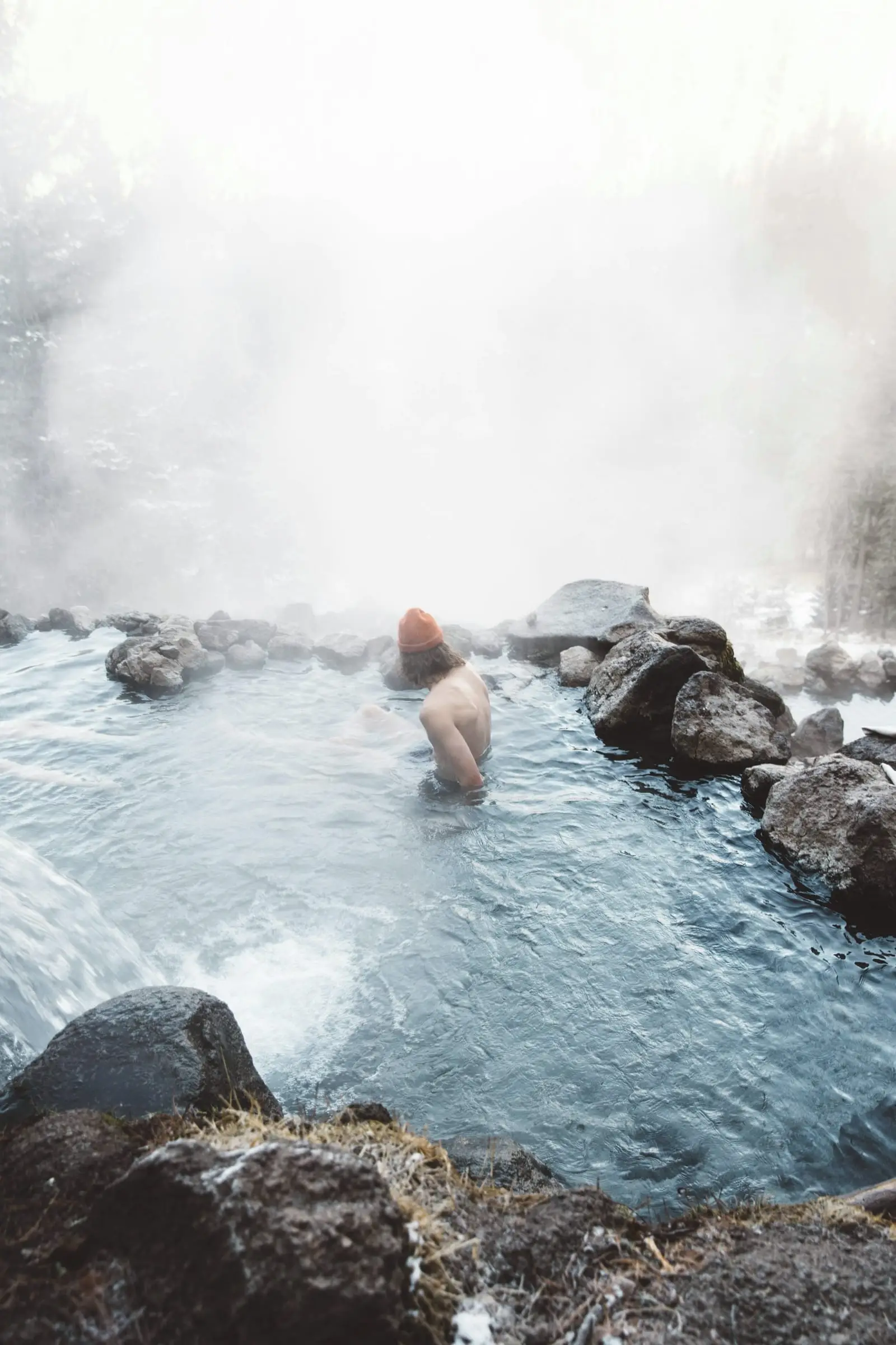 Una persona con gorro de lana sumergida en una poza de agua caliente entre rocas, envuelta en vapor.
