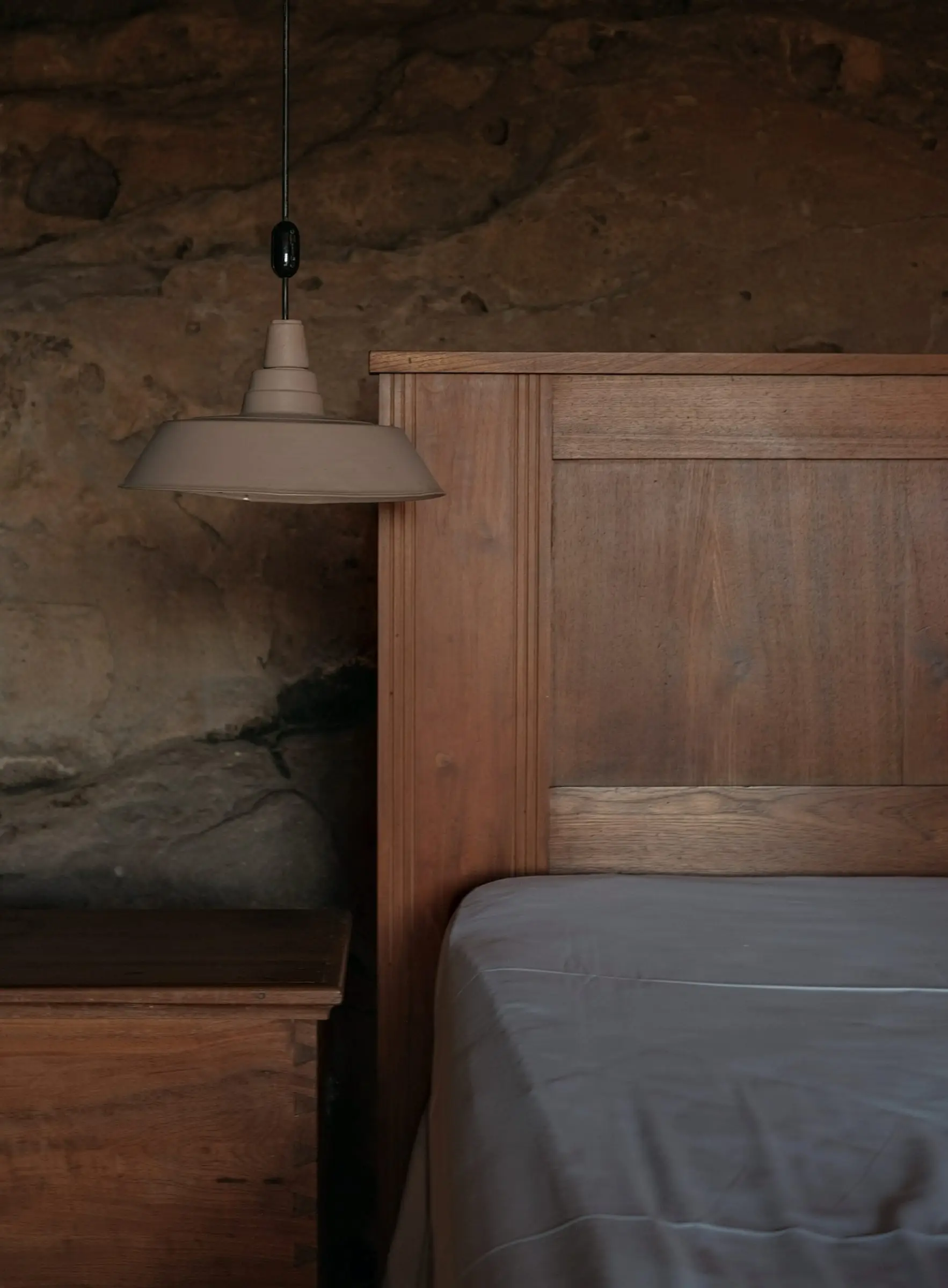 Cama con cabecero de madera encajada contra una pared de roca, iluminada por una lámpara colgante.