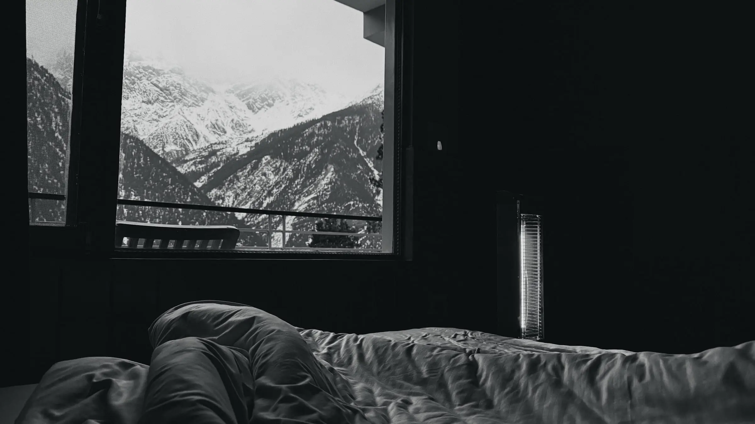 Cama deshecha junto a una ventana que da a montañas nevadas, en blanco y negro.