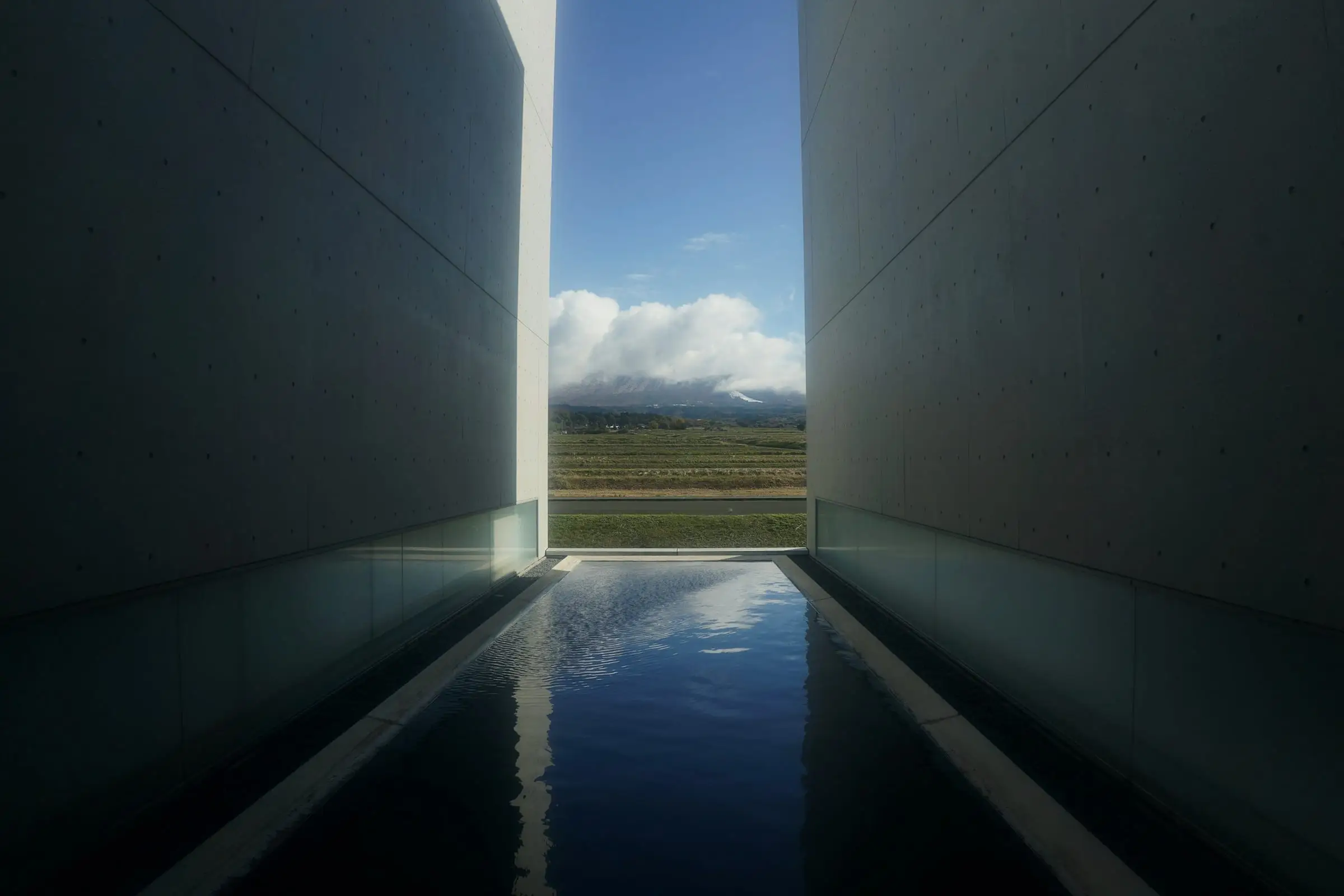 Un canal de agua quieta entre dos muros altos de hormigón; al fondo, por la abertura, una pradera y montañas bajo nubes.