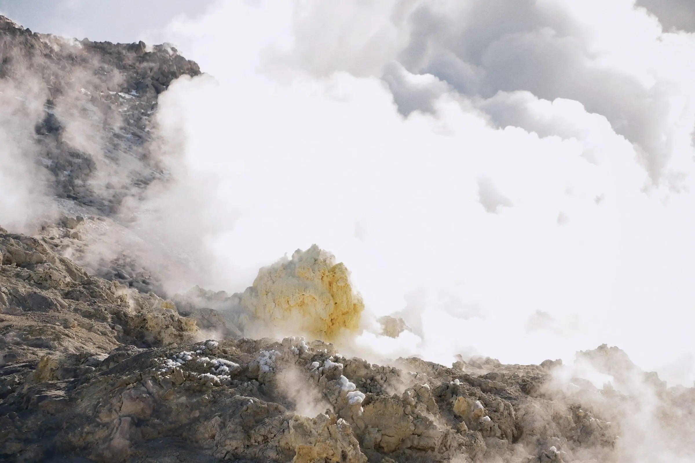 Fumarola entre rocas con depósitos amarillos de azufre y una nube densa de vapor blanco.