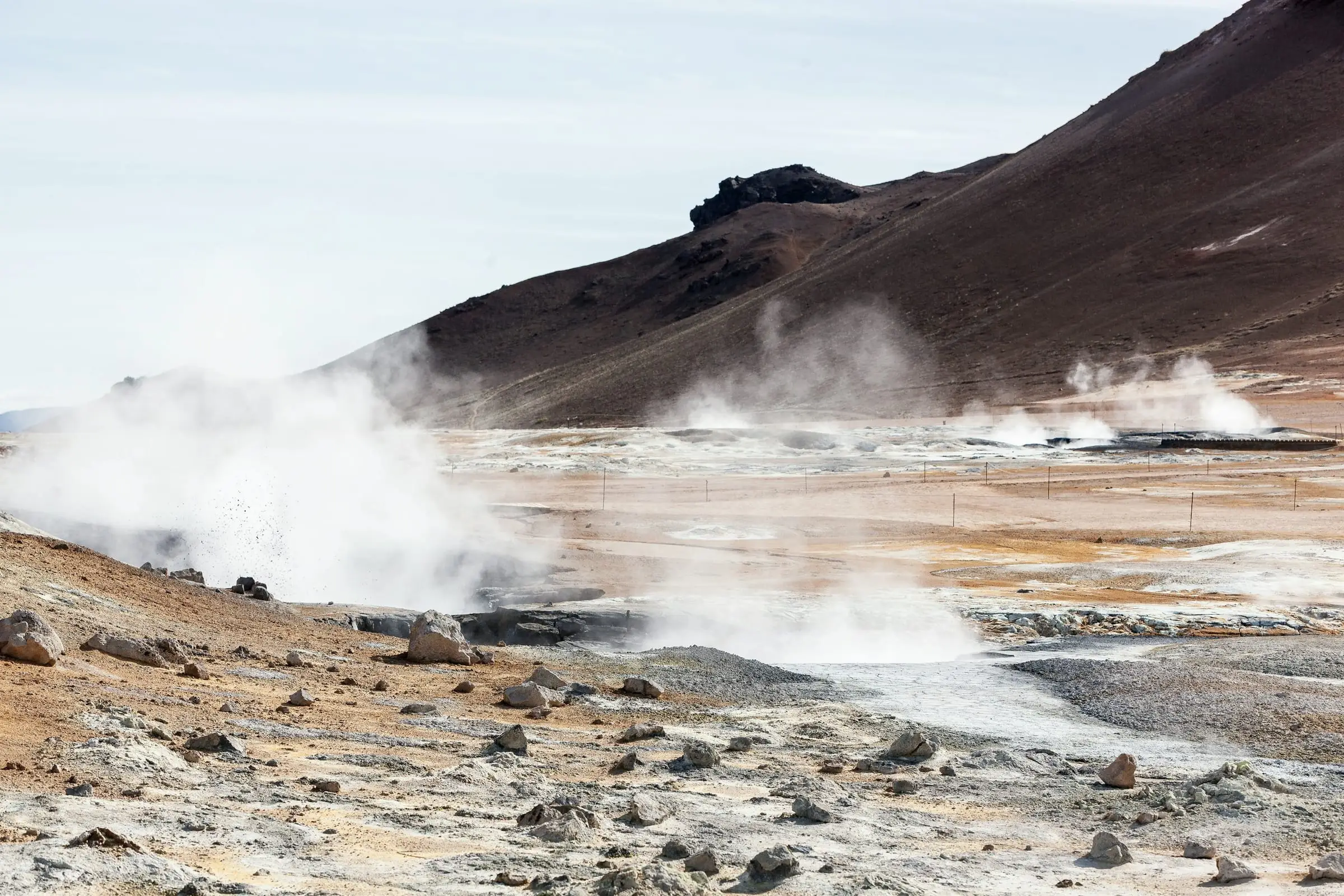 Llanura volcánica con columnas de vapor que salen del suelo, al pie de una montaña rojiza.