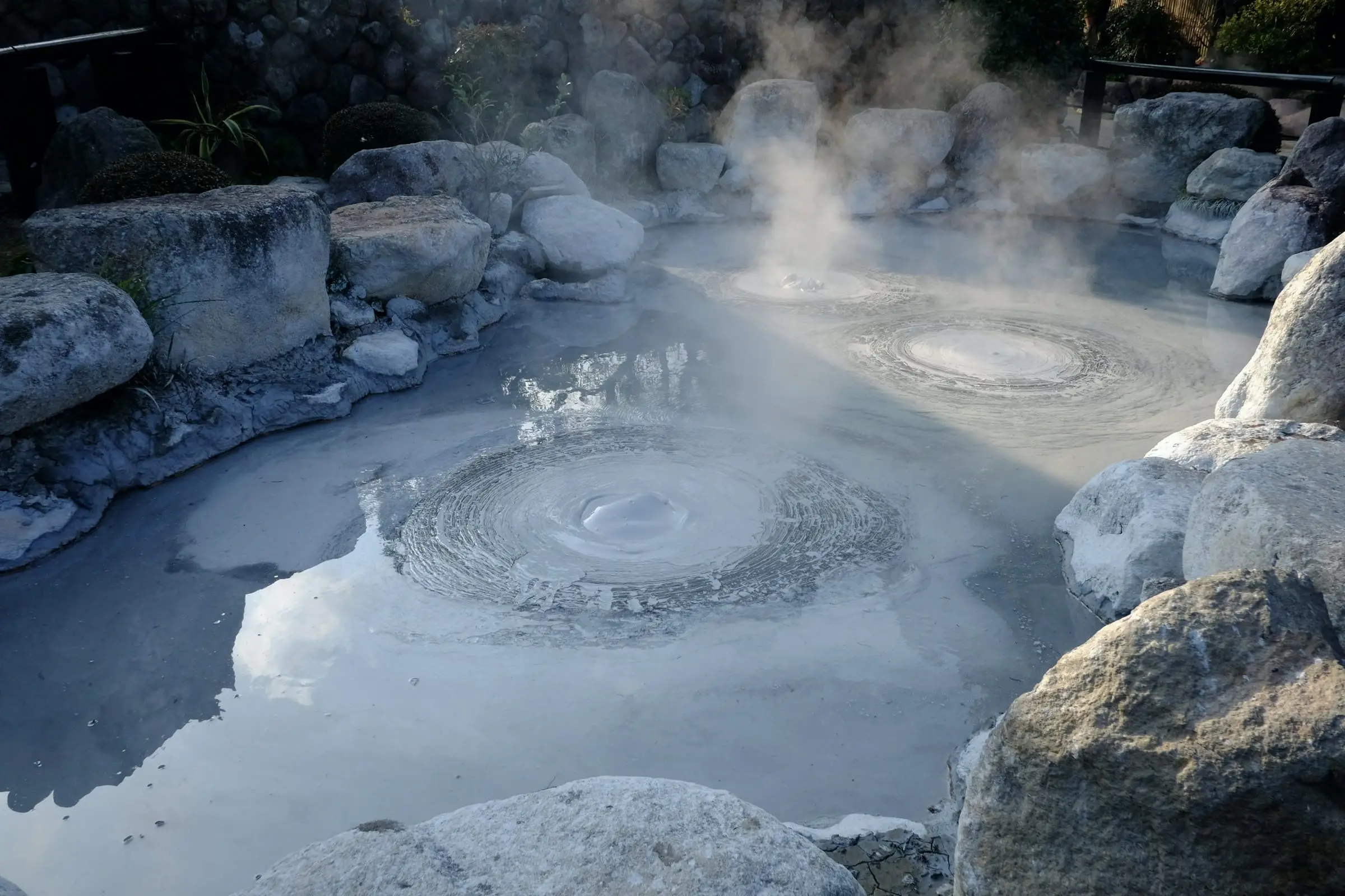 Un ojo de agua caliente entre rocas redondeadas: el agua burbujea en círculos concéntricos y suelta vapor.