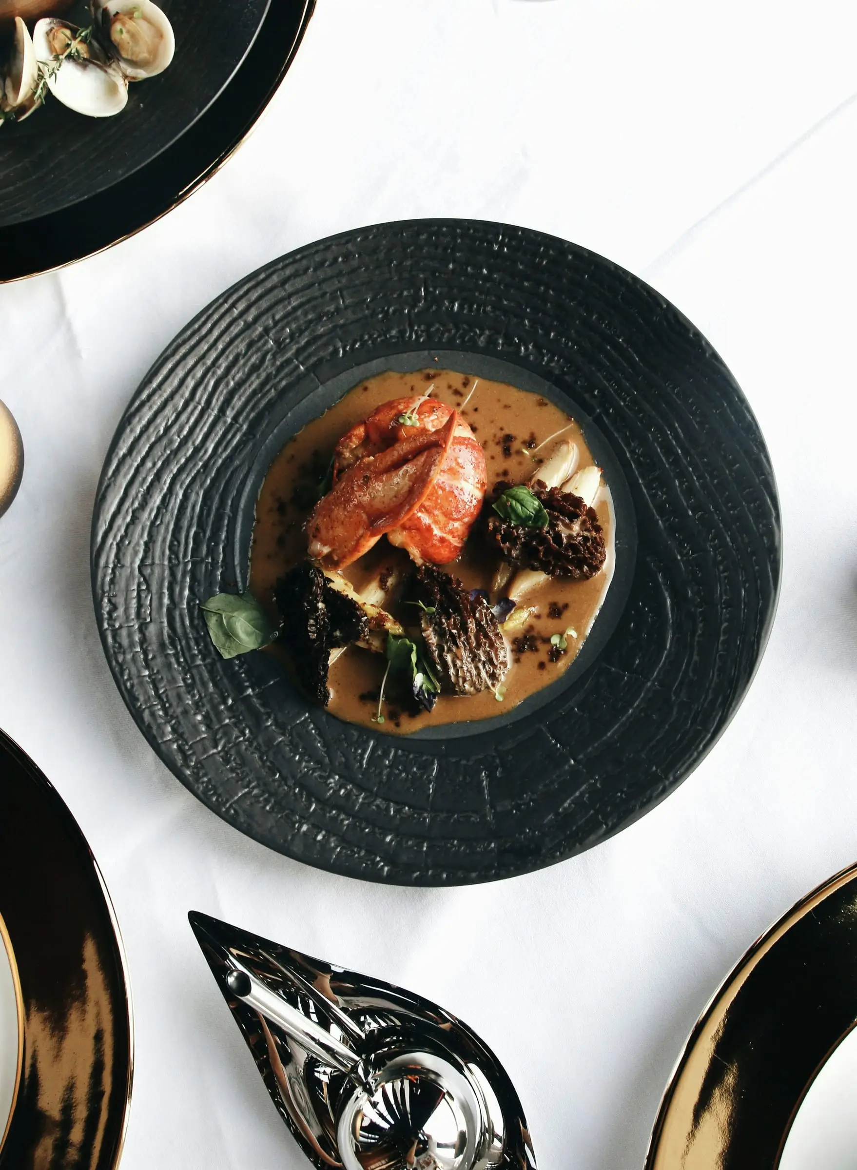 Plato de cerámica negra con trucha dorada, hongos y una salsa, visto desde arriba sobre mantel blanco.
