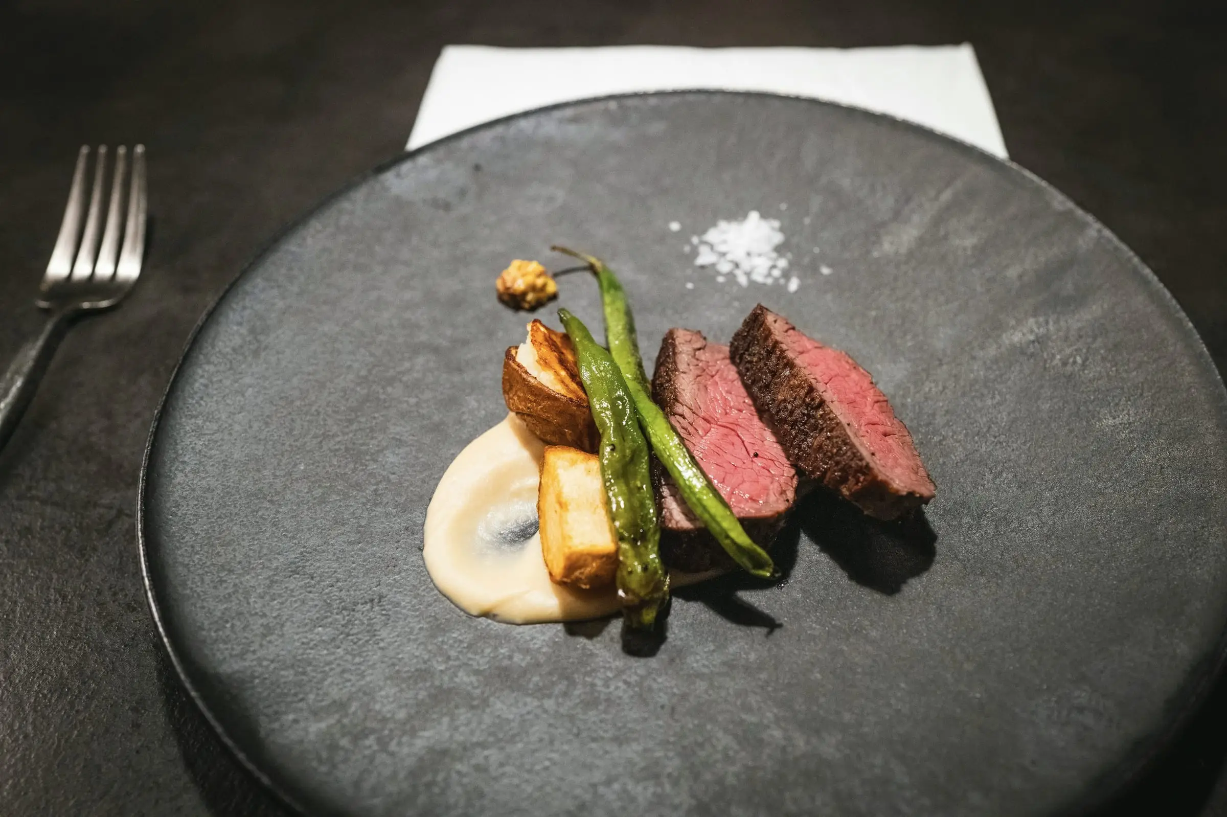 Carne roja cortada, chauchas y puré sobre un plato de piedra gris, en una mesa oscura.