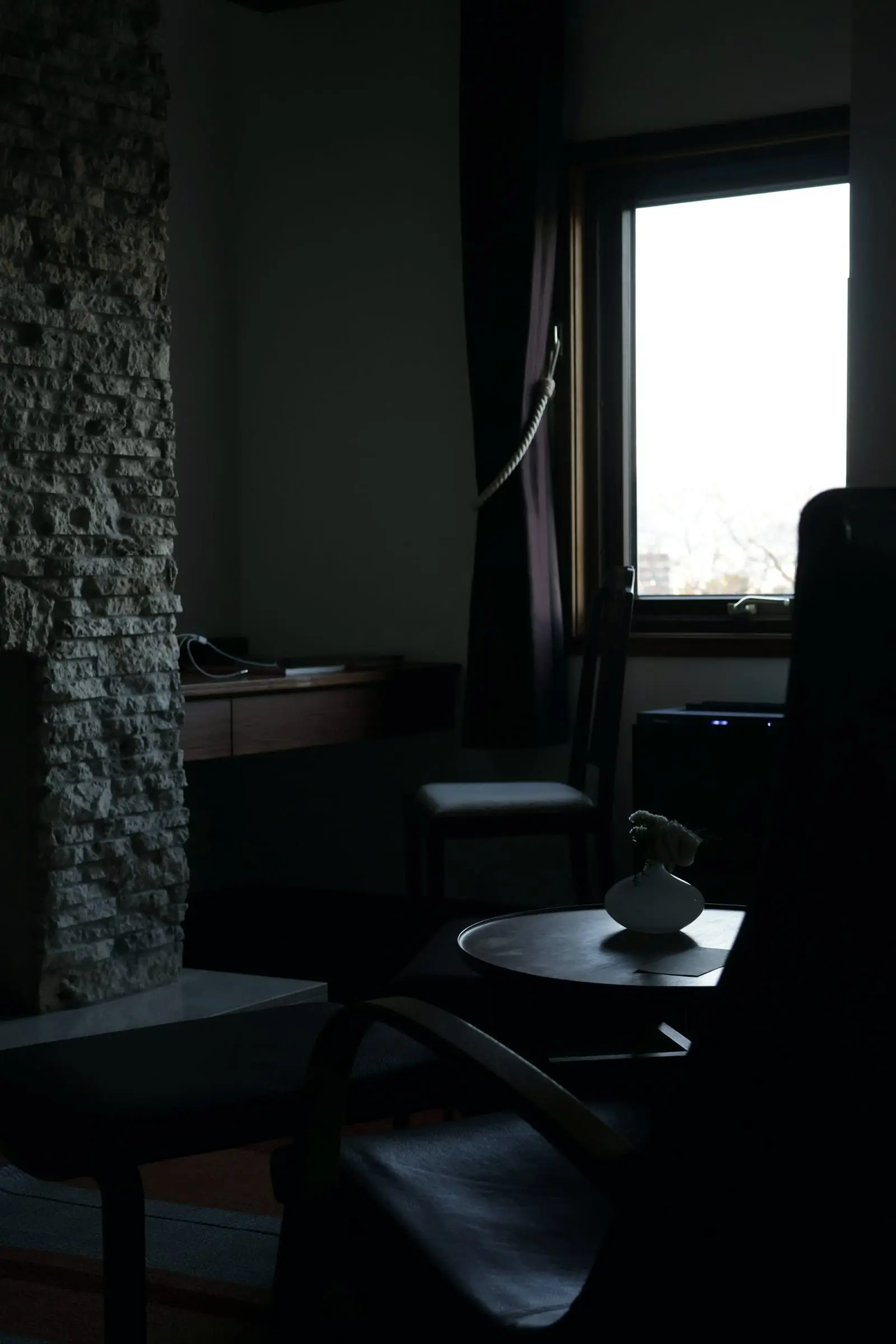 Rincón en penumbra con pared de piedra, un sillón y una mesa baja junto a una ventana.