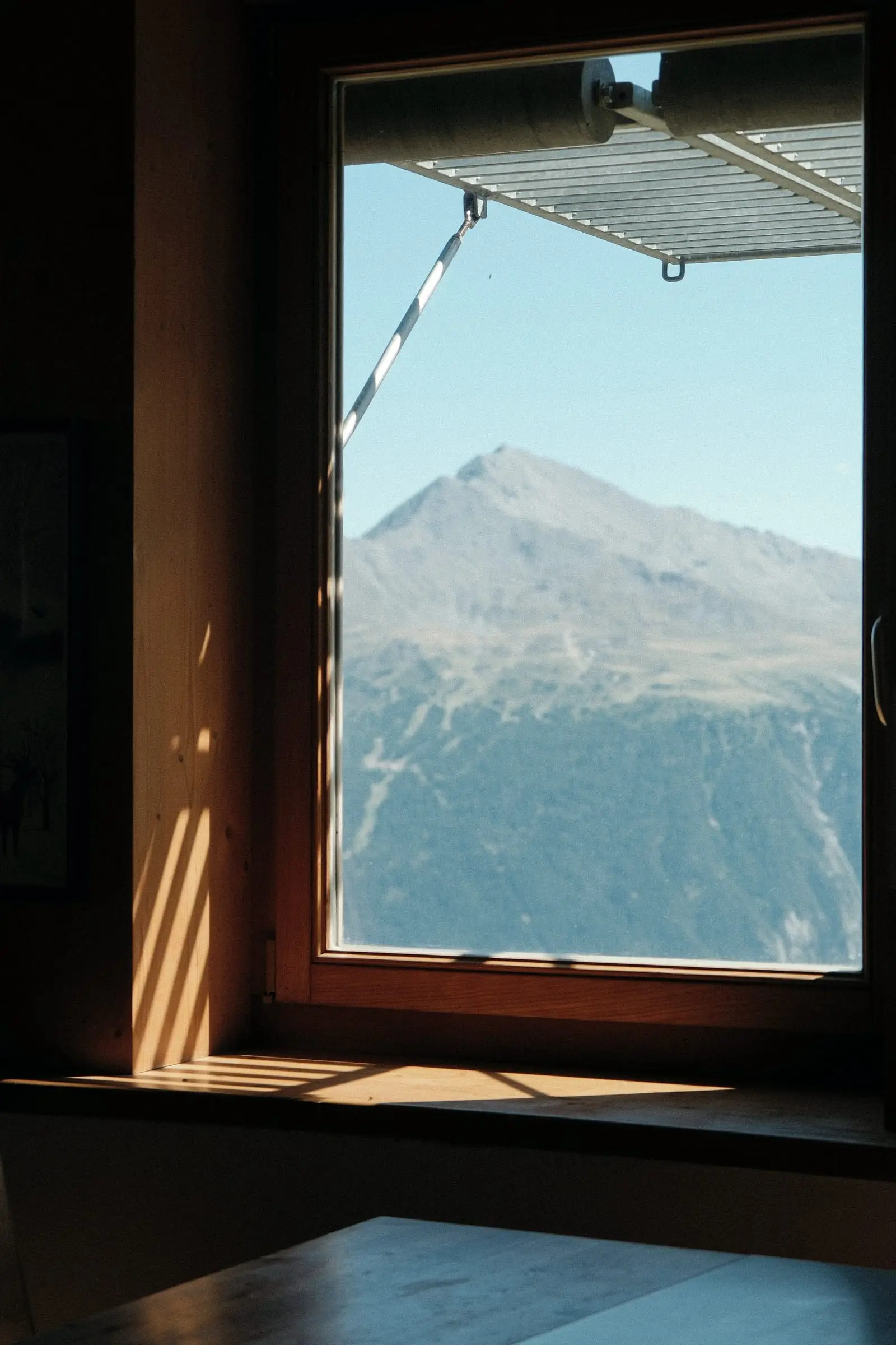 Ventana de madera abierta a una montaña con nieve; la luz de la tarde entra sobre el alféizar.