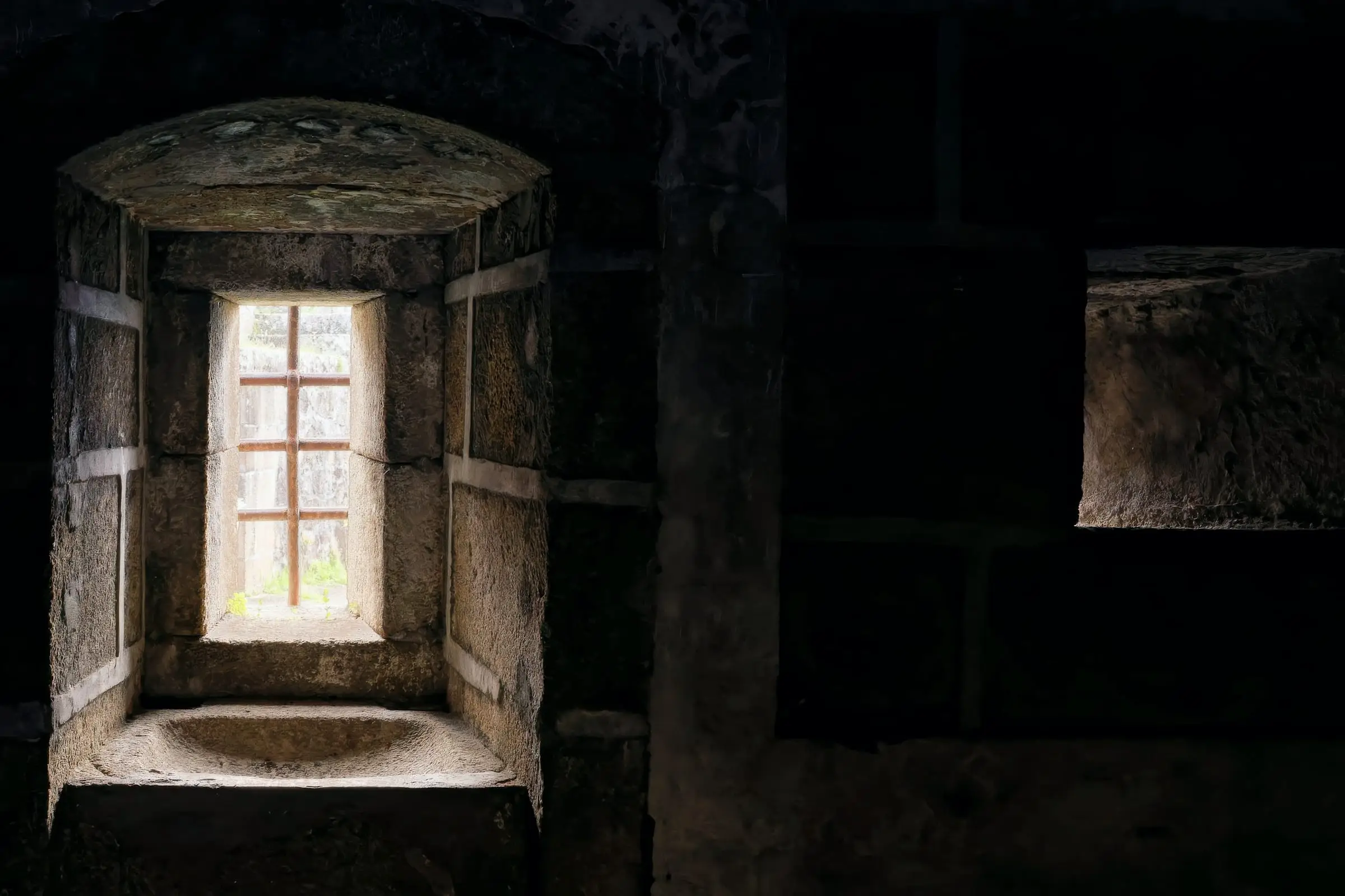 Ventana pequeña al fondo de un nicho de piedra con asiento, en un muro grueso y en penumbra.