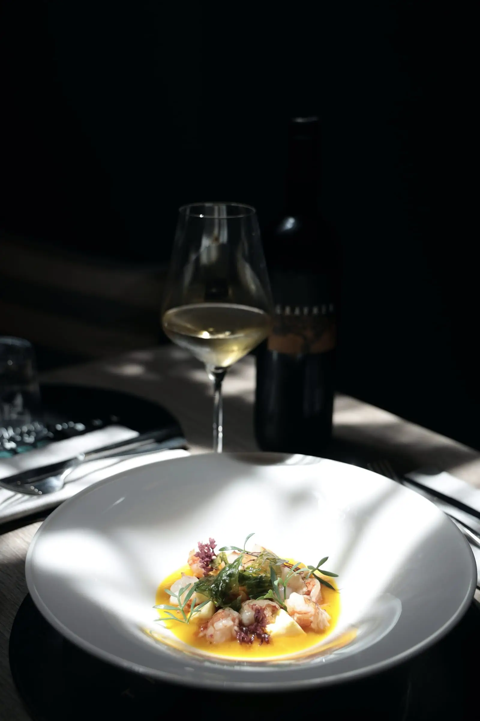 Copa de vino blanco y un plato con verduras sobre una mesa a contraluz.