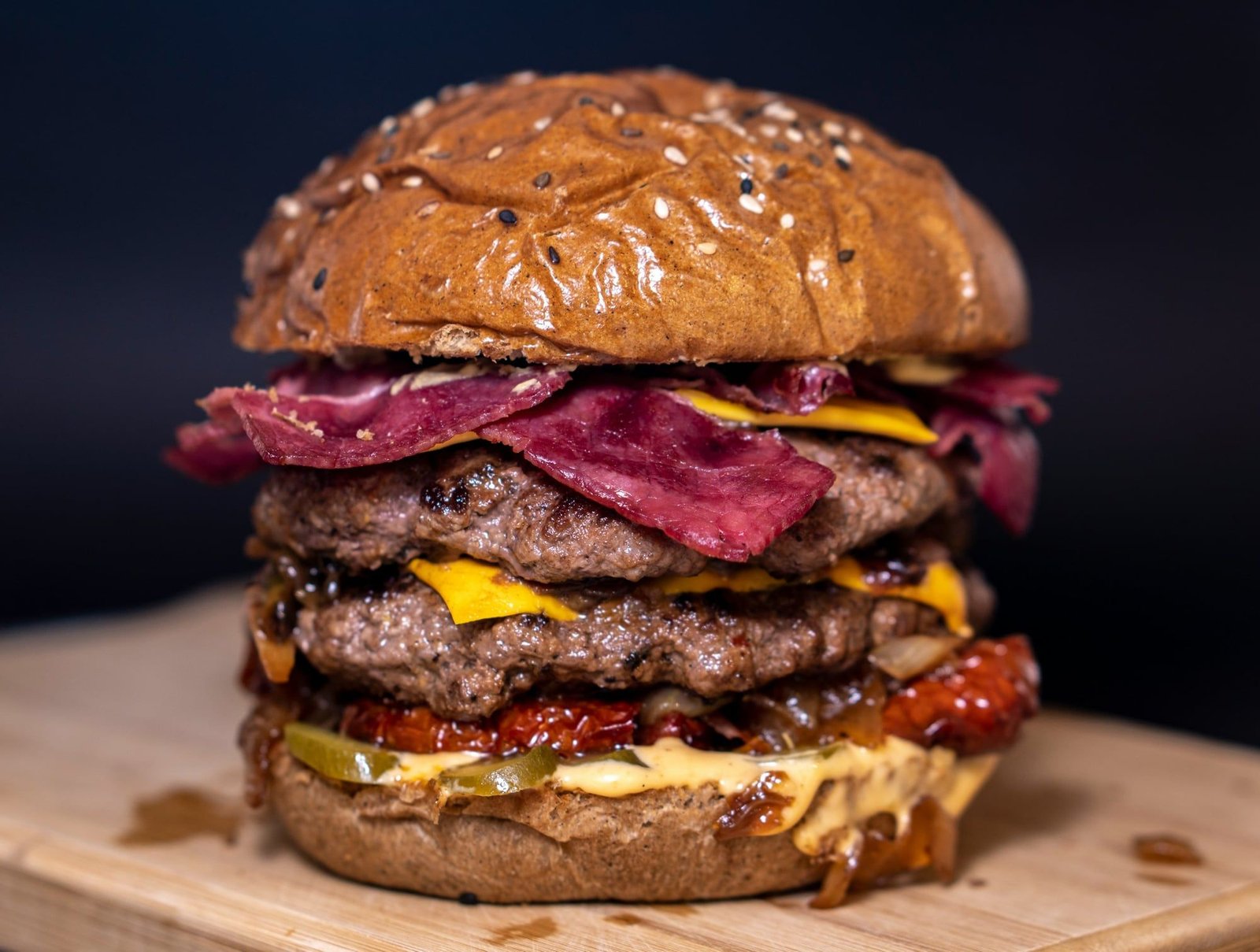 Hamburguesa doble con pastrami, cheddar, cebolla caramelizada y pepinos, sobre tabla de madera.