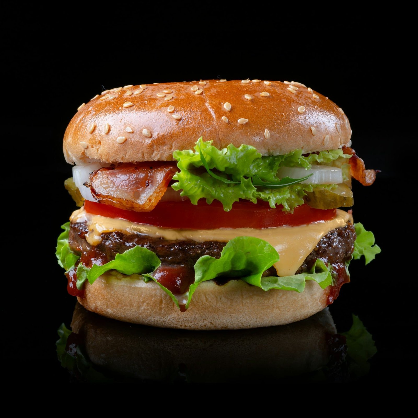 Hamburguesa alta con pan de sésamo, panceta, lechuga, tomate, cheddar y kétchup, sobre fondo negro con reflejo.