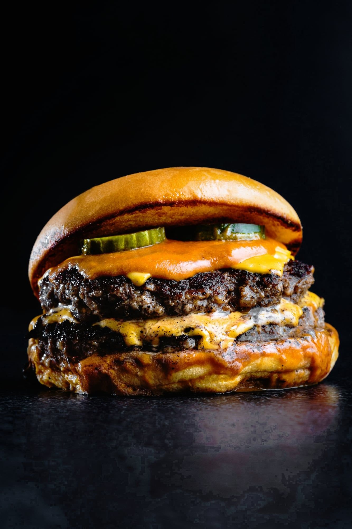 Hamburguesa doble con dos medallones de bordes crocantes, cheddar fundido, pepinos y salsa naranja, sobre fondo negro.