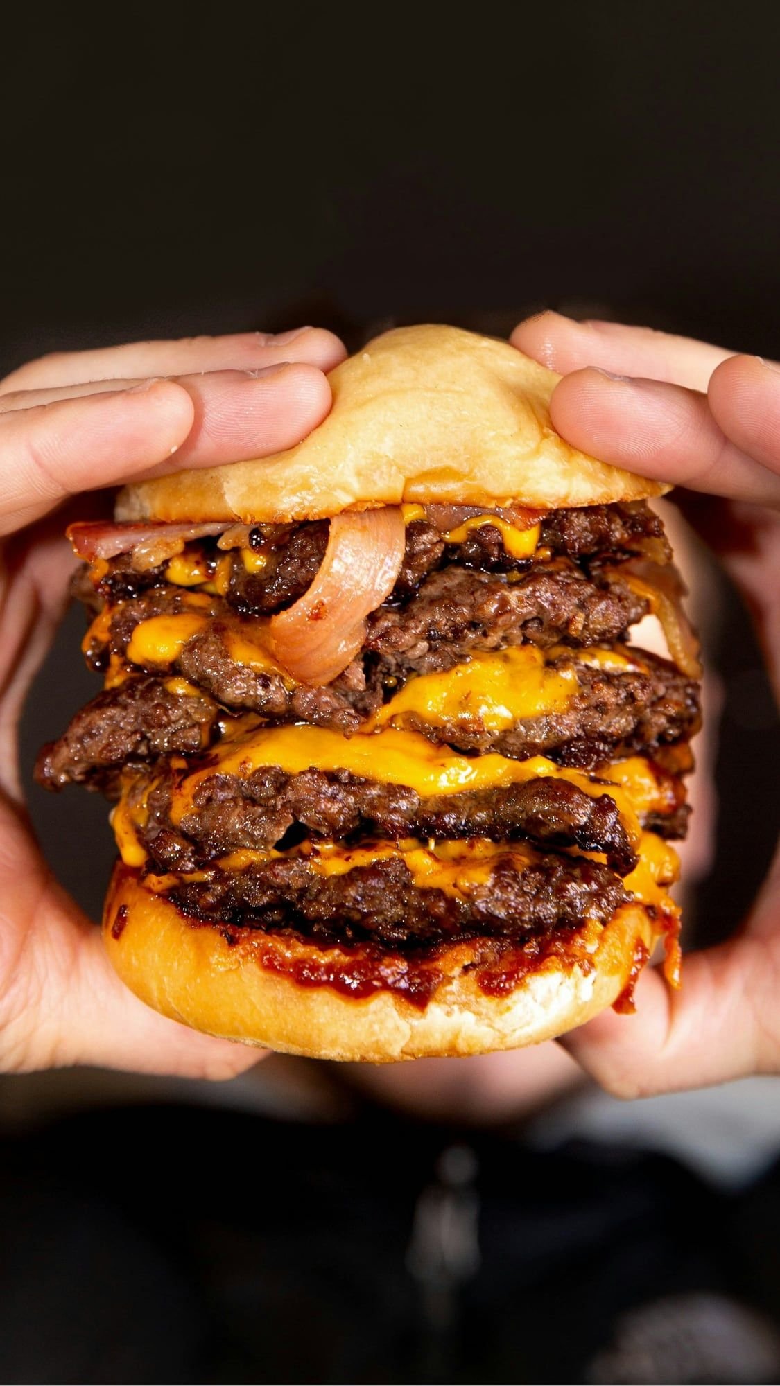 Dos manos aprietan una hamburguesa de varios pisos con cheddar fundido.