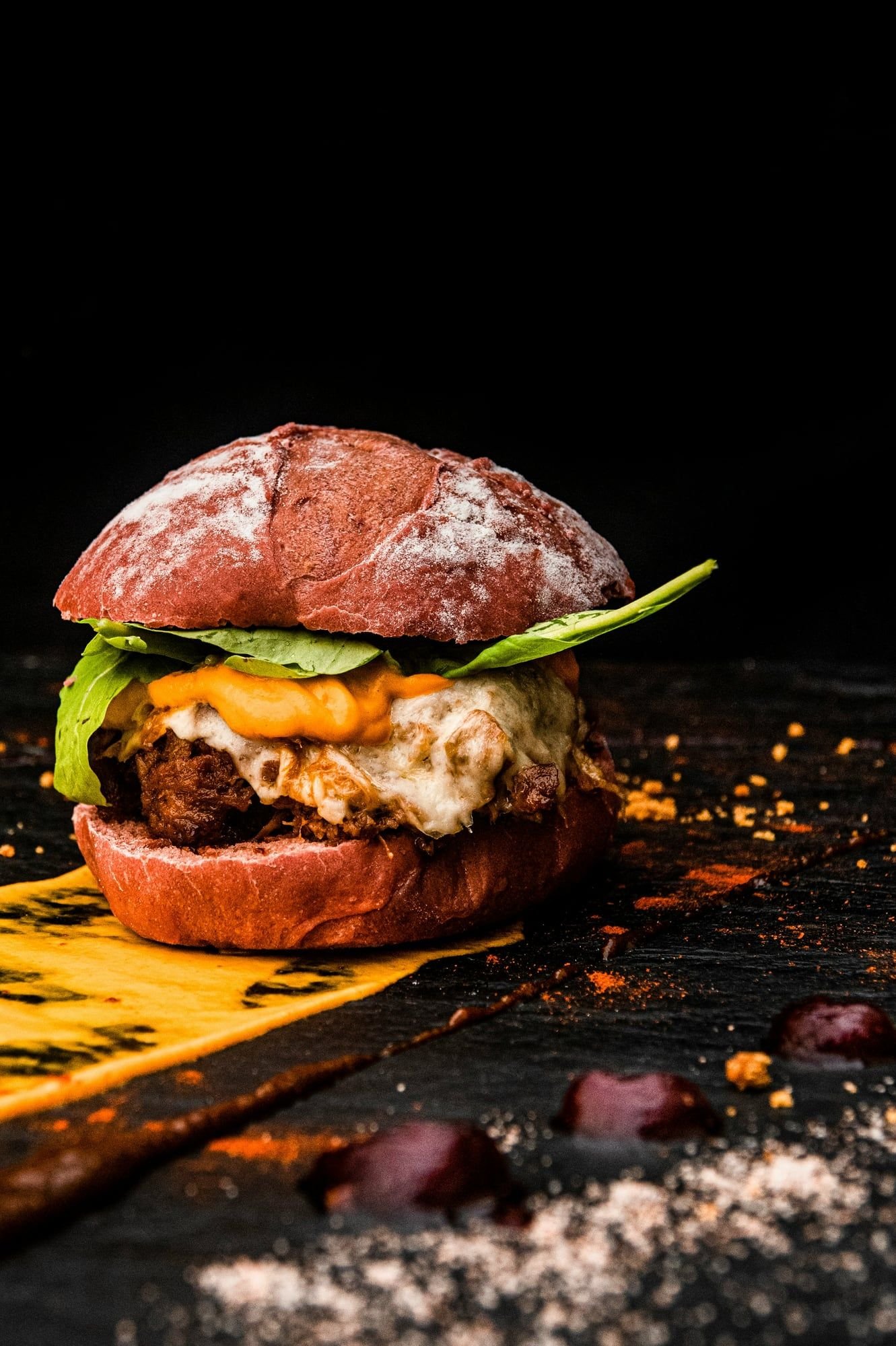 Hamburguesa con pan rojo enharinado, queso blanco fundido, salsa naranja y hojas verdes, sobre pizarra oscura con paprika.