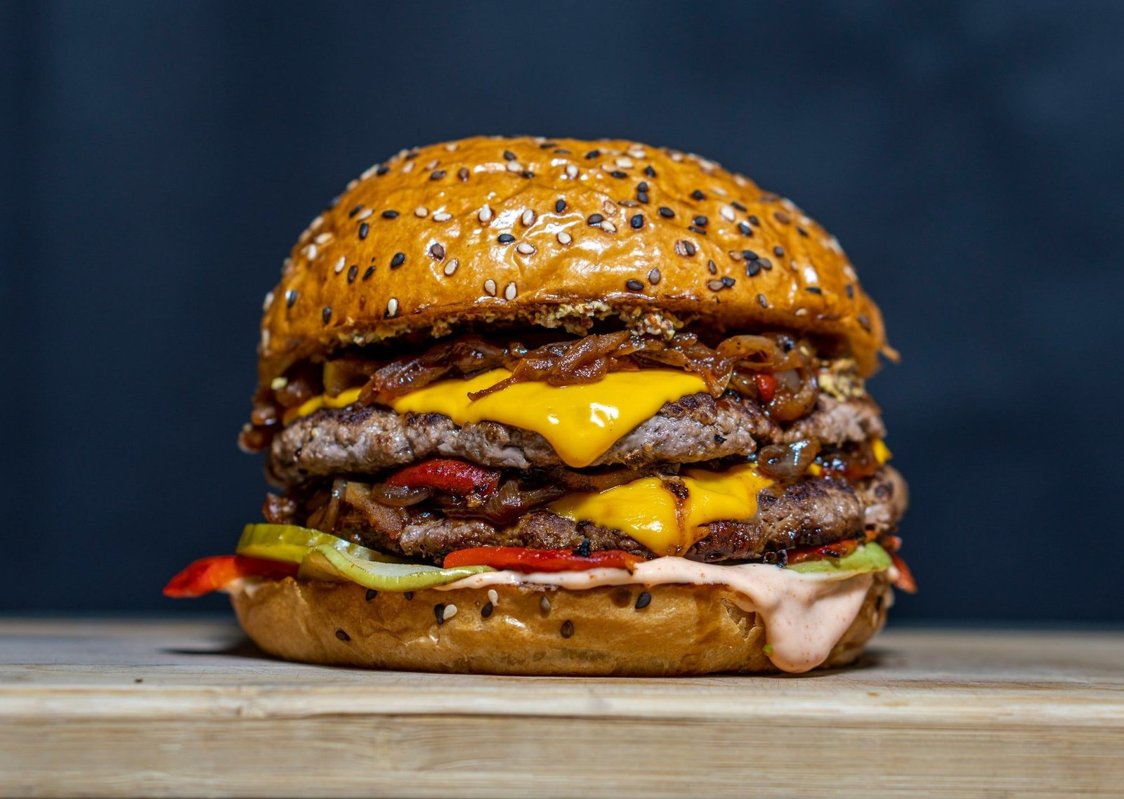 Hamburguesa doble con pan de sésamo blanco y negro, cebolla caramelizada, cheddar y morrones, sobre tabla de madera y fondo azul oscuro.