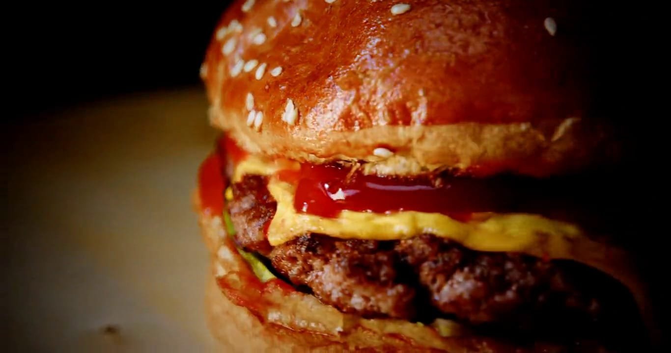 Hamburguesa doble con pan brioche brillante, cheddar y kétchup que gotea, girando sobre fondo oscuro.