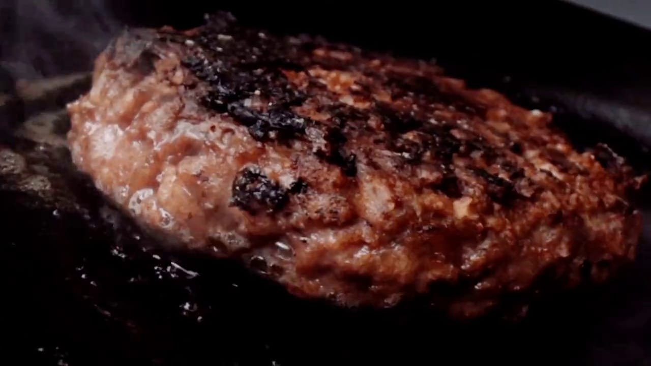 Primerísimo plano de un medallón sellándose sobre la plancha, con vapor; el hierro en blanco y negro, la carne a color.