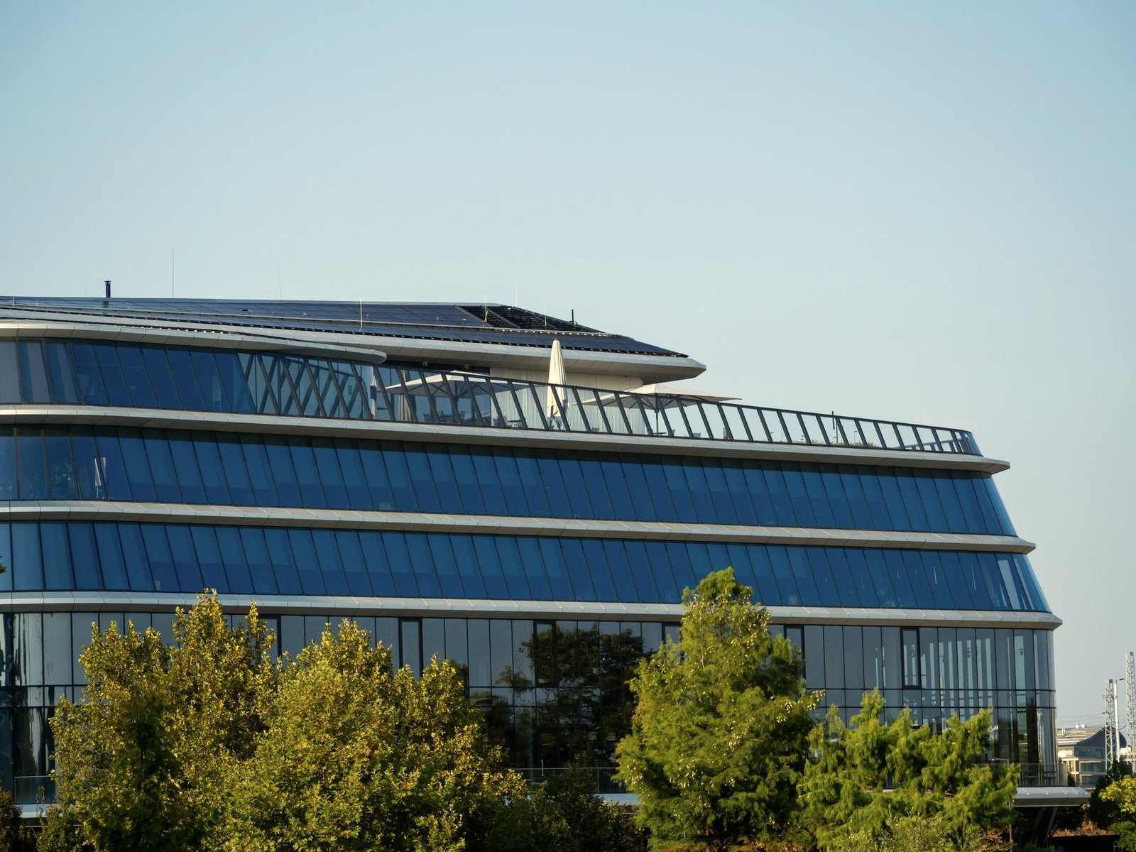 Edificio de oficinas de vidrio con plantas curvas y módulos solares en la cubierta, detrás de una línea de árboles.